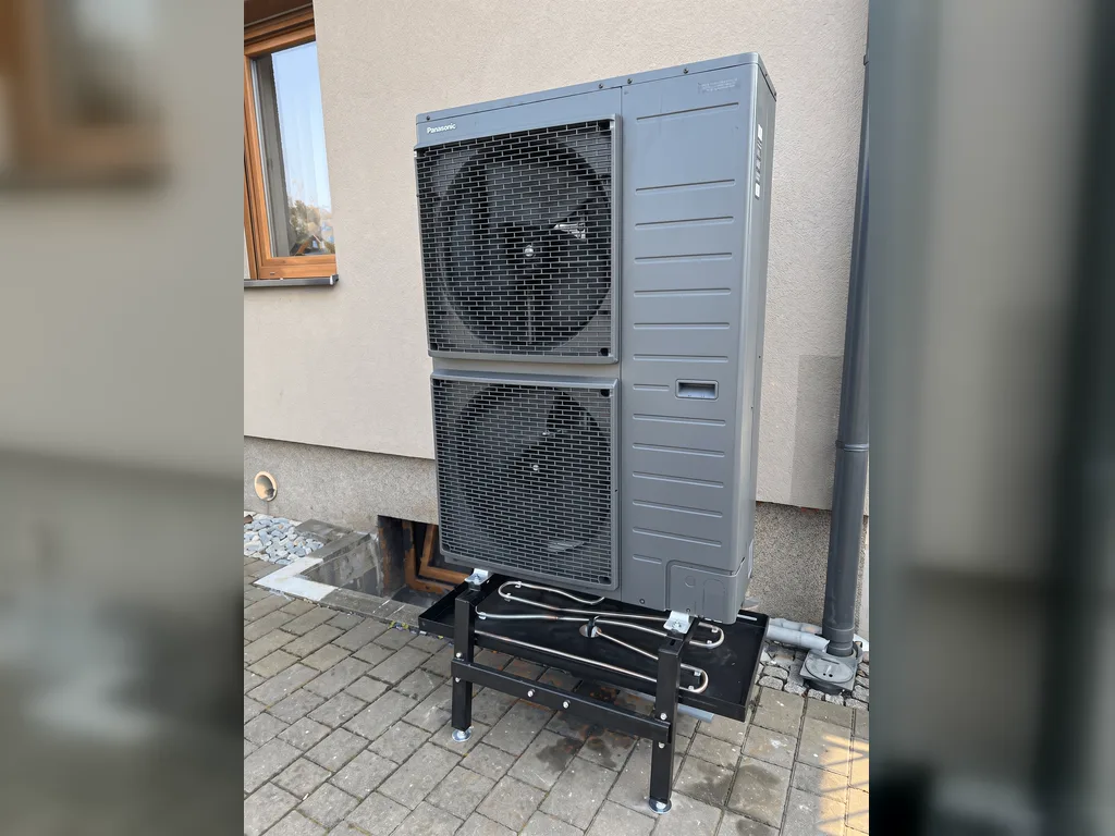 Ciemnoszara jednostka zewnętrzna pompy ciepła Panasonic z dwoma wentylatorami na stojaku przy ścianie domu