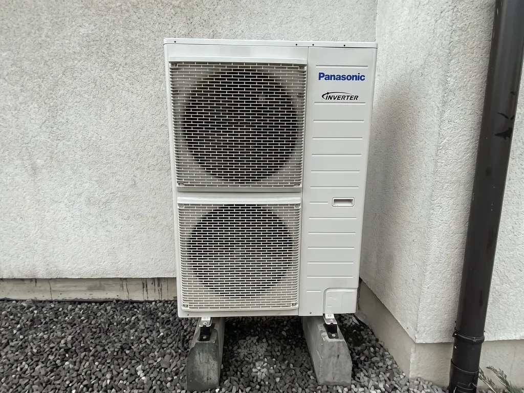 Pompa ciepła, Gorzyczki: Jednostka zewnętrzna Panasonic Inverter przy jasnej ścianie, żwir, rura spustowa