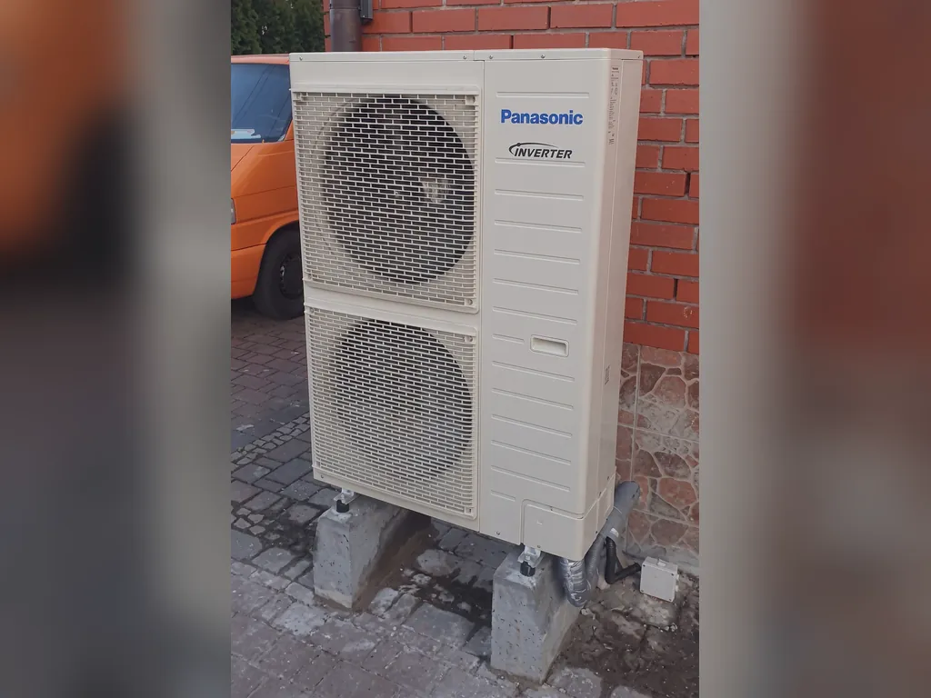 Pompa ciepła, Rybnik: Jednostka zewnętrzna Panasonic (dwa wentylatory) przy ceglanej ścianie; pionowe zdjęcie z białymi pasami po bokach