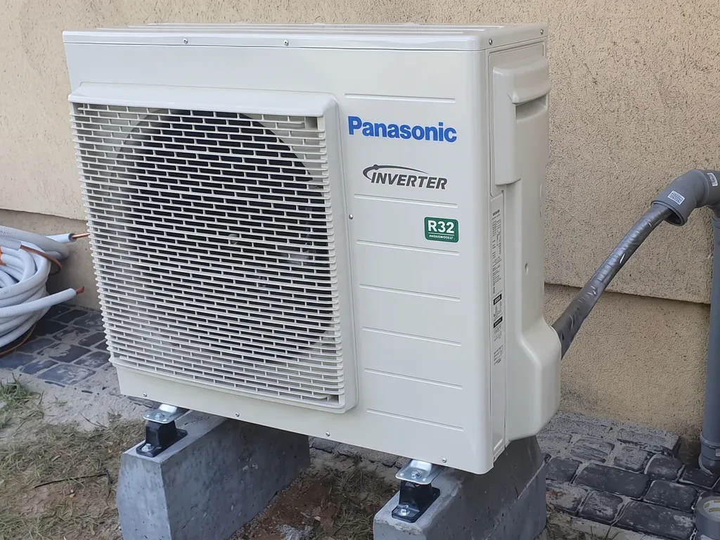 Pompa ciepła, Rybnik: Jednostka zewnętrzna Panasonic Inverter R32 (jeden wentylator) na bloczkach przy beżowej ścianie, rury