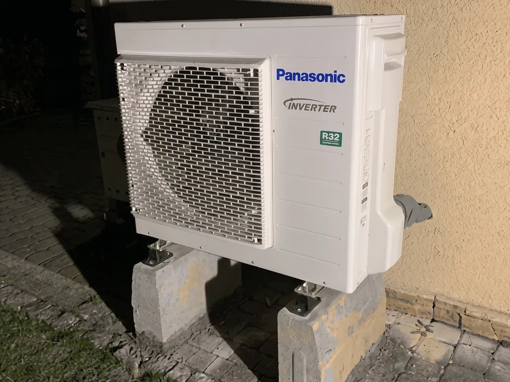 Pompa ciepła, Orzesze: Jednostka zewnętrzna Panasonic R32 na bloczkach przy beżowej ścianie, nocą z lampą błyskową