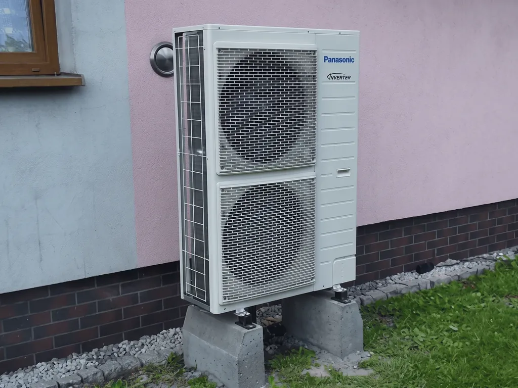 Pompa ciepła, Rybnik: Jednostka zewnętrzna Panasonic (dwa wentylatory) na bloczkach przy różowo-białej ścianie z ceglanym cokołem, trawnik