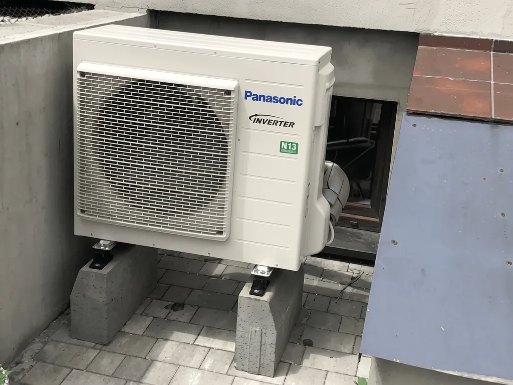 Pompa ciepła, Pogrzebień: Jednostka zewnętrzna Panasonic Inverter R32 na bloczkach przy szarej ścianie z płytą