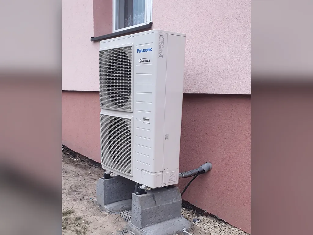 Pompa ciepła, Krostoszowice: Jednostka zewnętrzna Panasonic przy różowo-brązowej ścianie, piasek; białe pasy