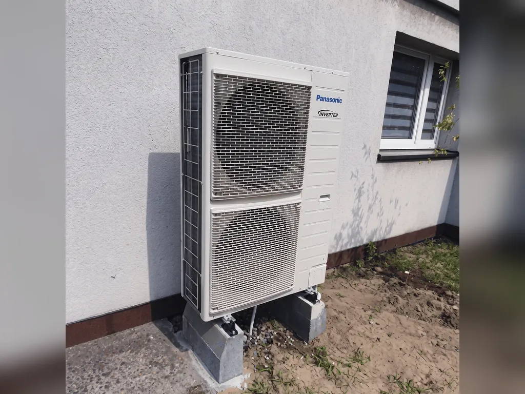 Pompa ciepła, Rybnik: Jednostka zewnętrzna Panasonic na bloczkach przy białej ścianie, piasek; białe pasy