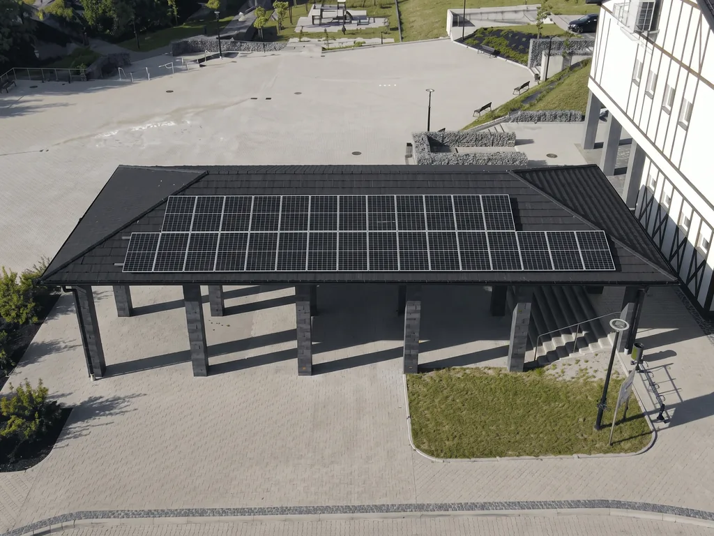 Wiata na słupach z panelami fotowoltaicznymi na dachu (carport) na placu przy budynku z muru pruskiego, widok z drona